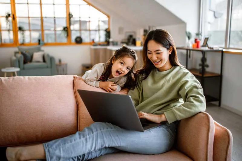 Woman on a couch with her young daughter, using her laptop to choose the best tax filing status for her taxes.