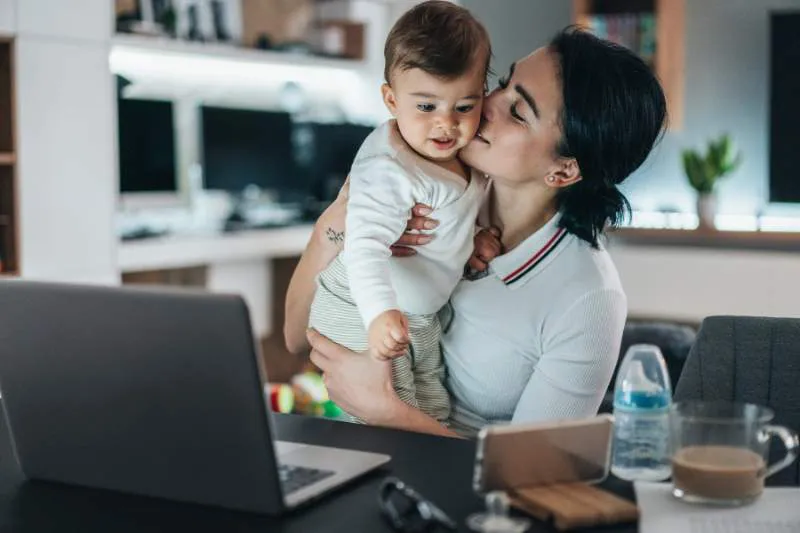 A mother kisses her baby while working at her laptop, highlighting the importance of family in understanding dependents for tax purposes.